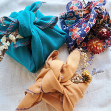 Load image into Gallery viewer, Three eco friendly fabric gift wraps tied in furoshiki style, one turquoise, one orange, one blue patterned. All with dried flower posy