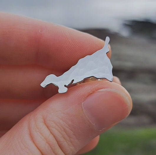Kernow Cornwall map shaped silver brooch pin held in fingers, turned to show hammered texture, Portscatho beach in background