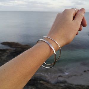 Arm and hand wearing silver rings and two recycled silver wave bangles, over backdrop of Cornish waves and beach