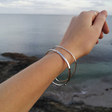Load image into Gallery viewer, Arm wearing silver ripple and wild wave bangles around wrist, against Cornish beach backdrop