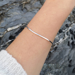 Close up of sustainable recycled silver curved wavy bangle, worn on arm with white jumper sleeve against grey rocks