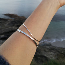 Load image into Gallery viewer, Close up of arm wearing two silver wave stacking bangles, against background of Cornish beach and ocean
