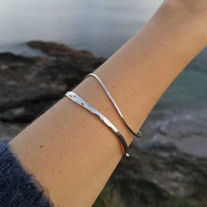 Close up of arm wearing larger and smaller silver wave bangles, sea and rocks in background