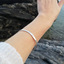 Load image into Gallery viewer, Silver wave bangle worn on arm with white jumper sleeve, in front of Cornish sea and rocks
