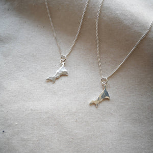 Two Lowen Cornwall map necklaces hanging from recycled silver chains on plain calico fabric