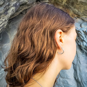 Handmade Cornish wave splash earrings worn on beach with slate cliffs in Cornwall, side view