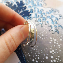 Load image into Gallery viewer, Thumb and finger holding silver wild wave hoop earrings, showing hammered texture on front, with wave in background
