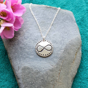Personalised recycled silver love token coin necklace, engraved with infinity symbol, on stone background with pink flowers