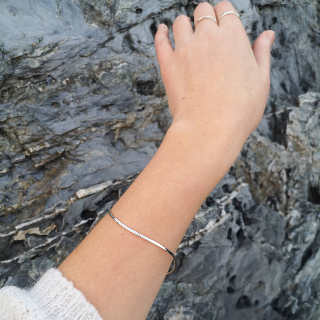 Silver minimalist curved bangle, worn on woman's left arm with background of grey cornish rocks