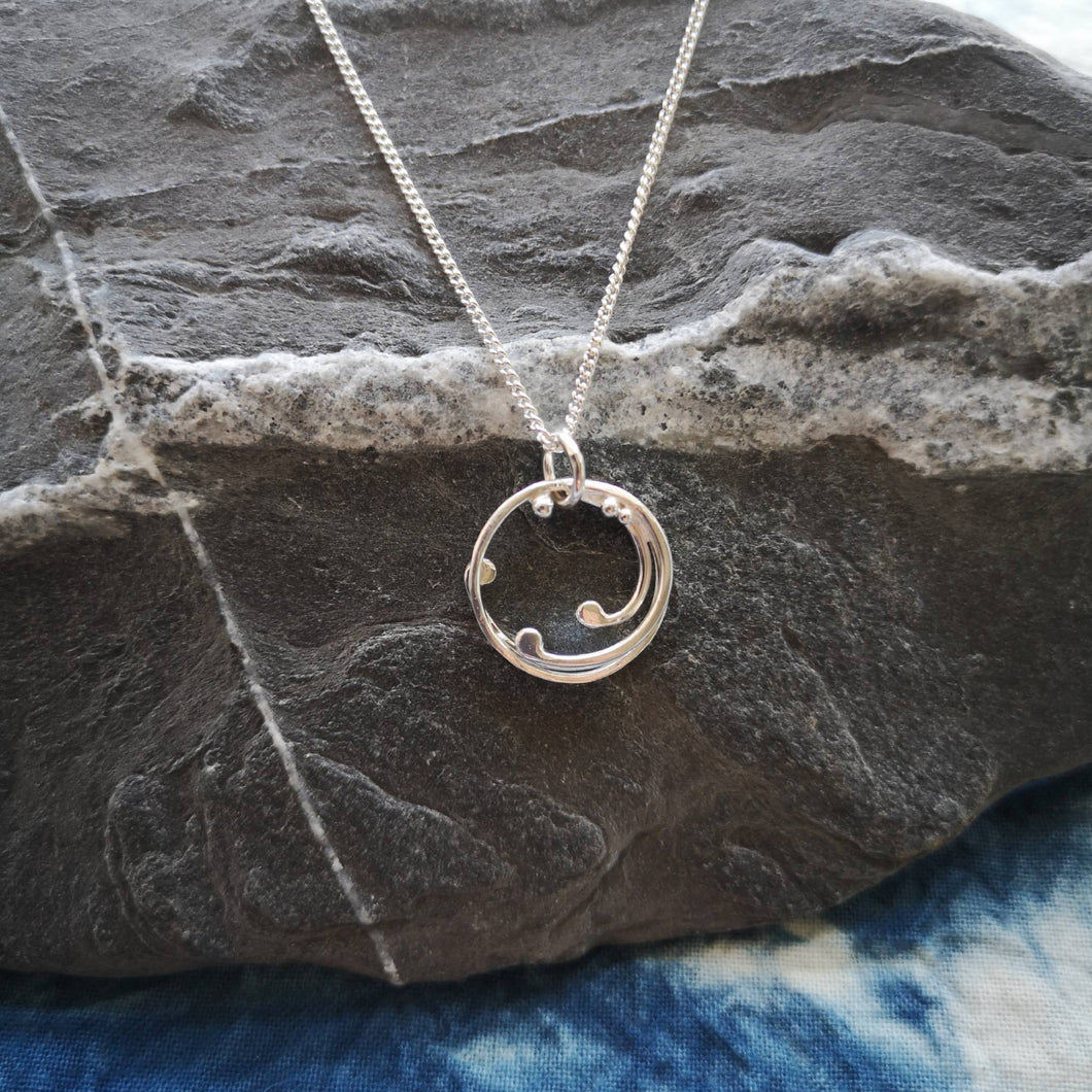 Cornish wave recycled silver necklace hung from grey and and white stone, on blue and white fabric
