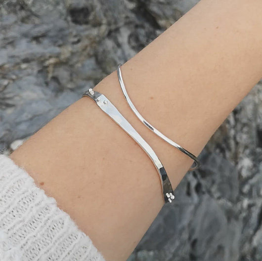 Arm wearing two handmade silver wave bangles, turning slightly to show the texture and reflection, against rocks 